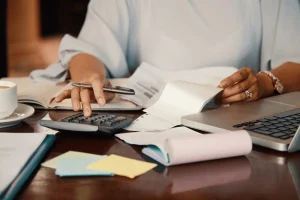 An professional calculates finances using a calculator, laptop, and paperwork, with coffee and sticky notes on a wooden desk.