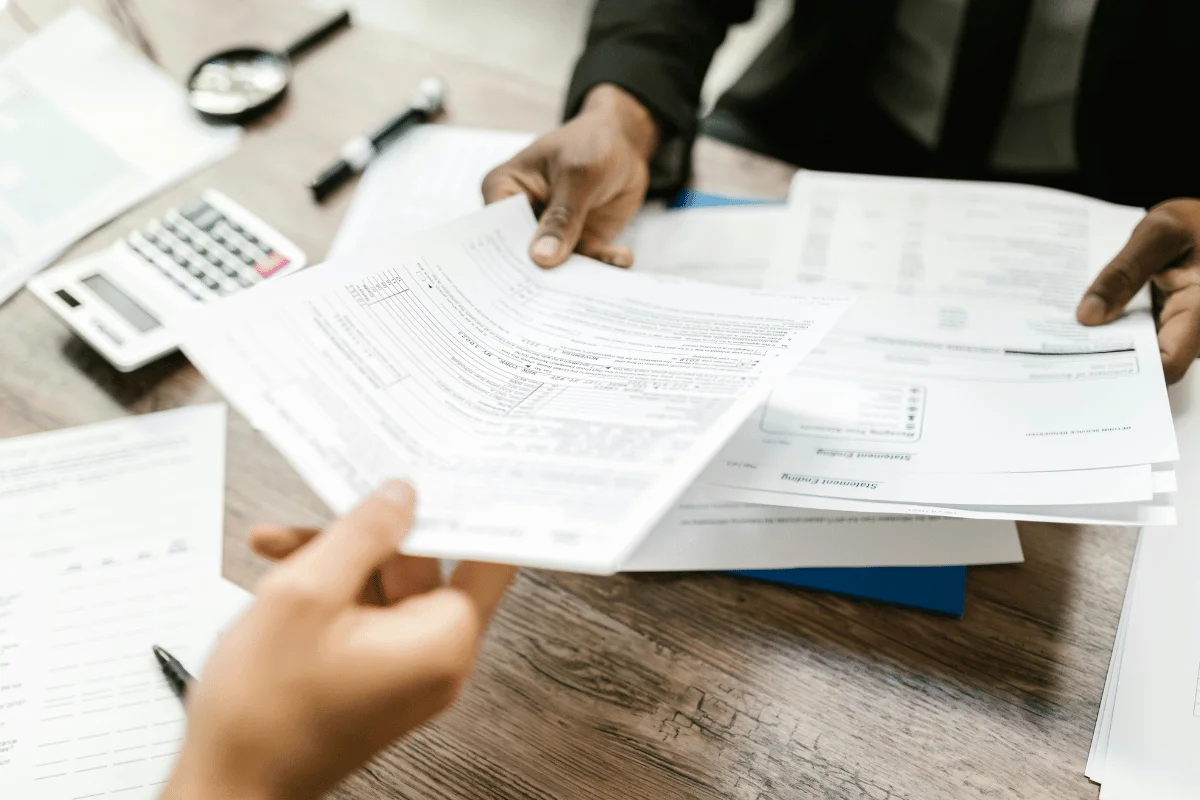 Two individuals exchange documents over a wooden desk with financial forms, a calculator, and pens. The tone suggests a professional business setting.