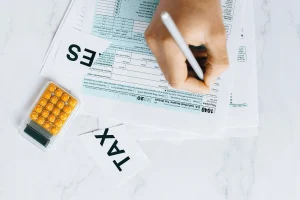 A hand writes on tax forms while a calculator with yellow buttons and a "TAX" label are nearby.