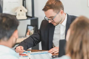 A person in a suit points to financial charts and data on a table while engaged in a discussion with two others.