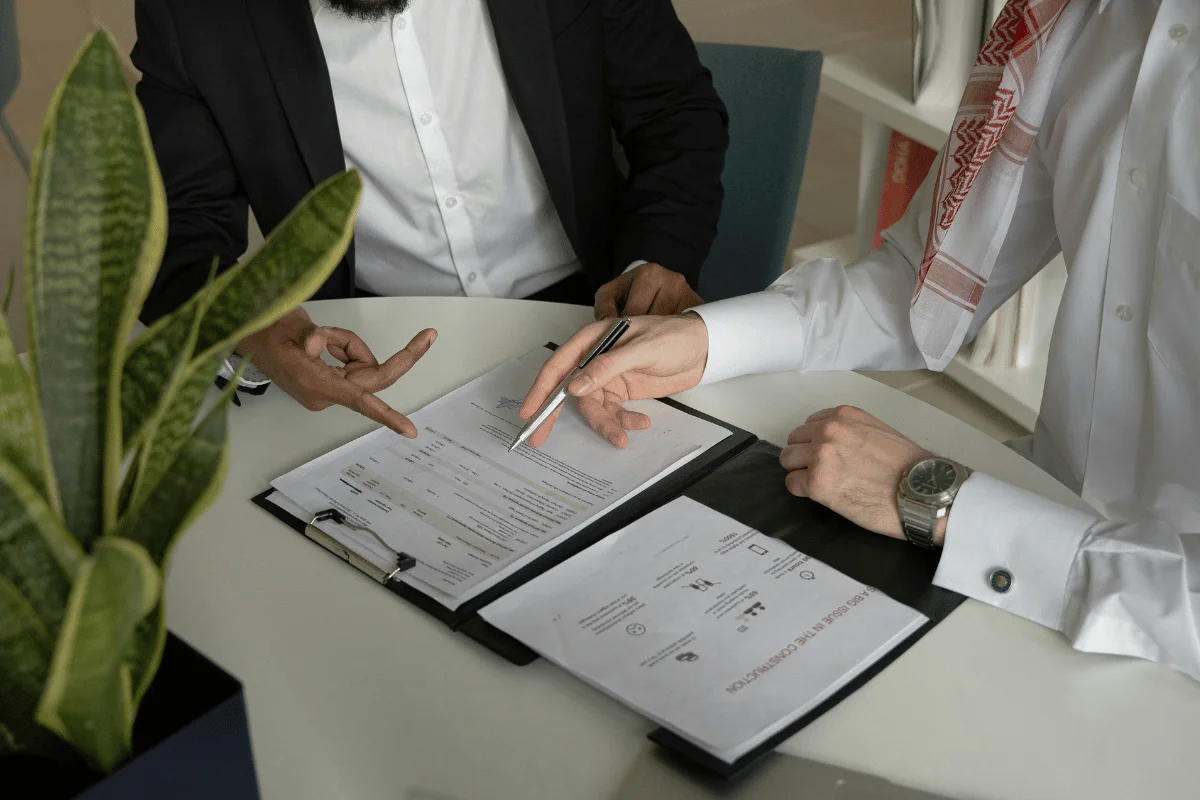 Two men in business attire discussing documents at a table, with one pointing and the other holding a pen, beside a leafy plant.
