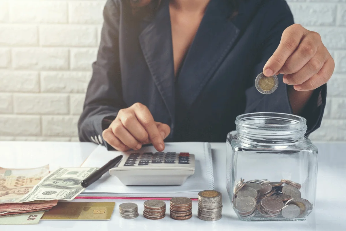 A woman places a coin into a jar while working on her finances with money and a calculator on the table.