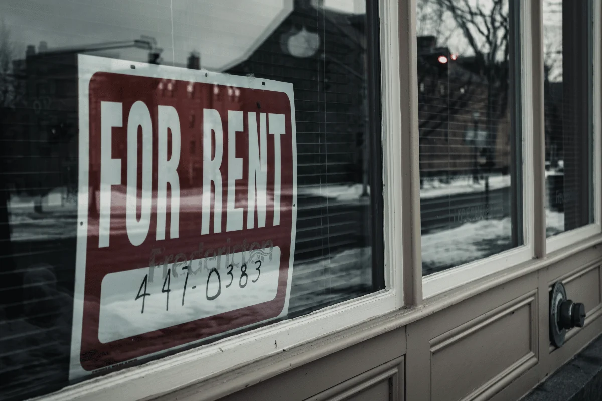 A "For Rent" sign in a window, featuring a contact number, with a snowy street view reflected behind it.