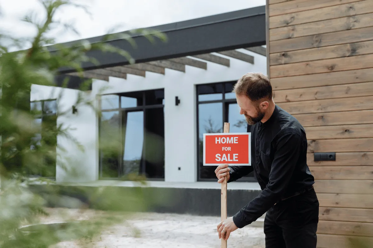 A man standing outside a modern home, holding a "Home for Sale" sign.