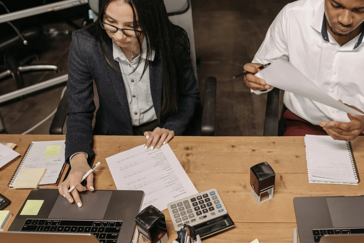 Two individuals collaborate at a wooden desk with a laptop, papers, a calculator, and stationery, engaged in a productive meeting.
