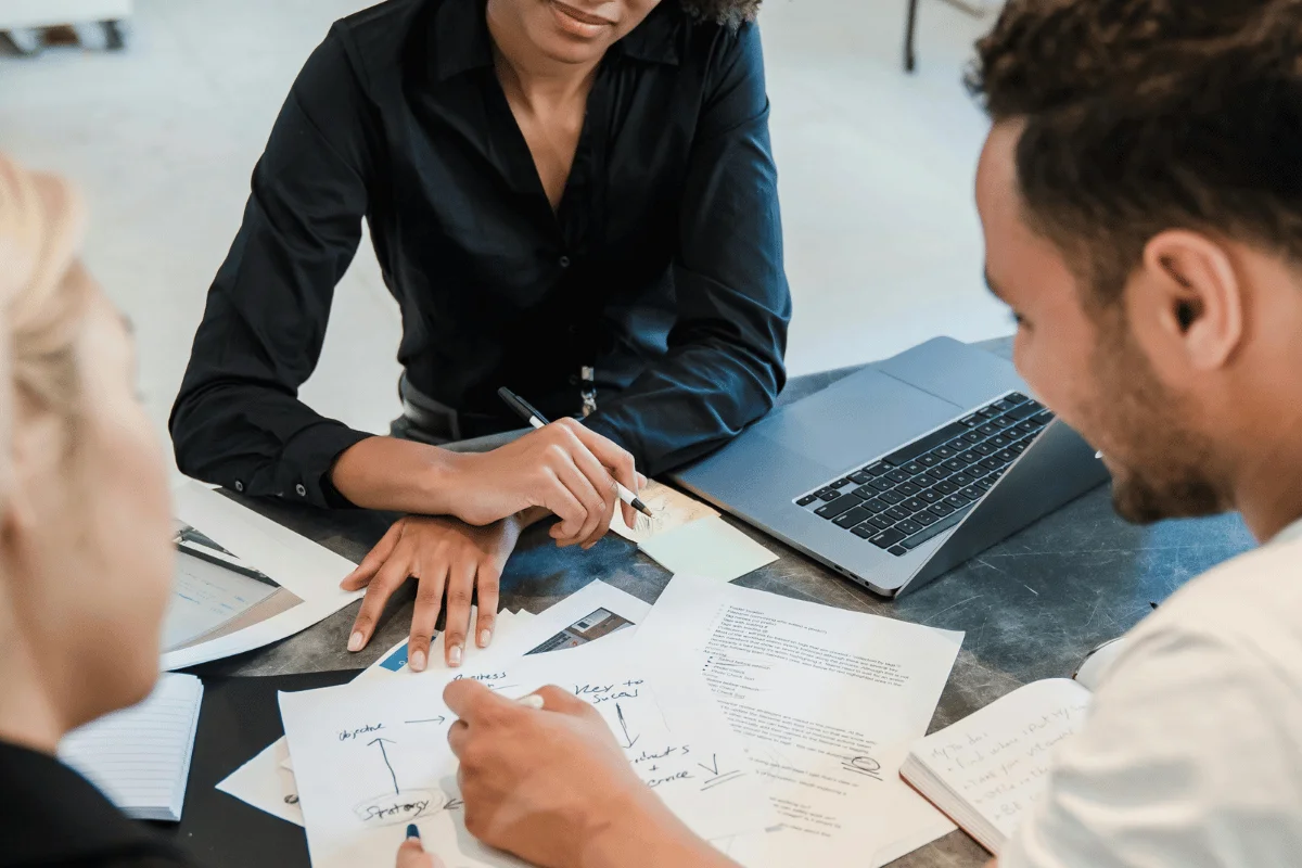 Three colleagues collaborate around a table covered with documents, notes, and a laptop. Their expressions suggest focus and teamwork.