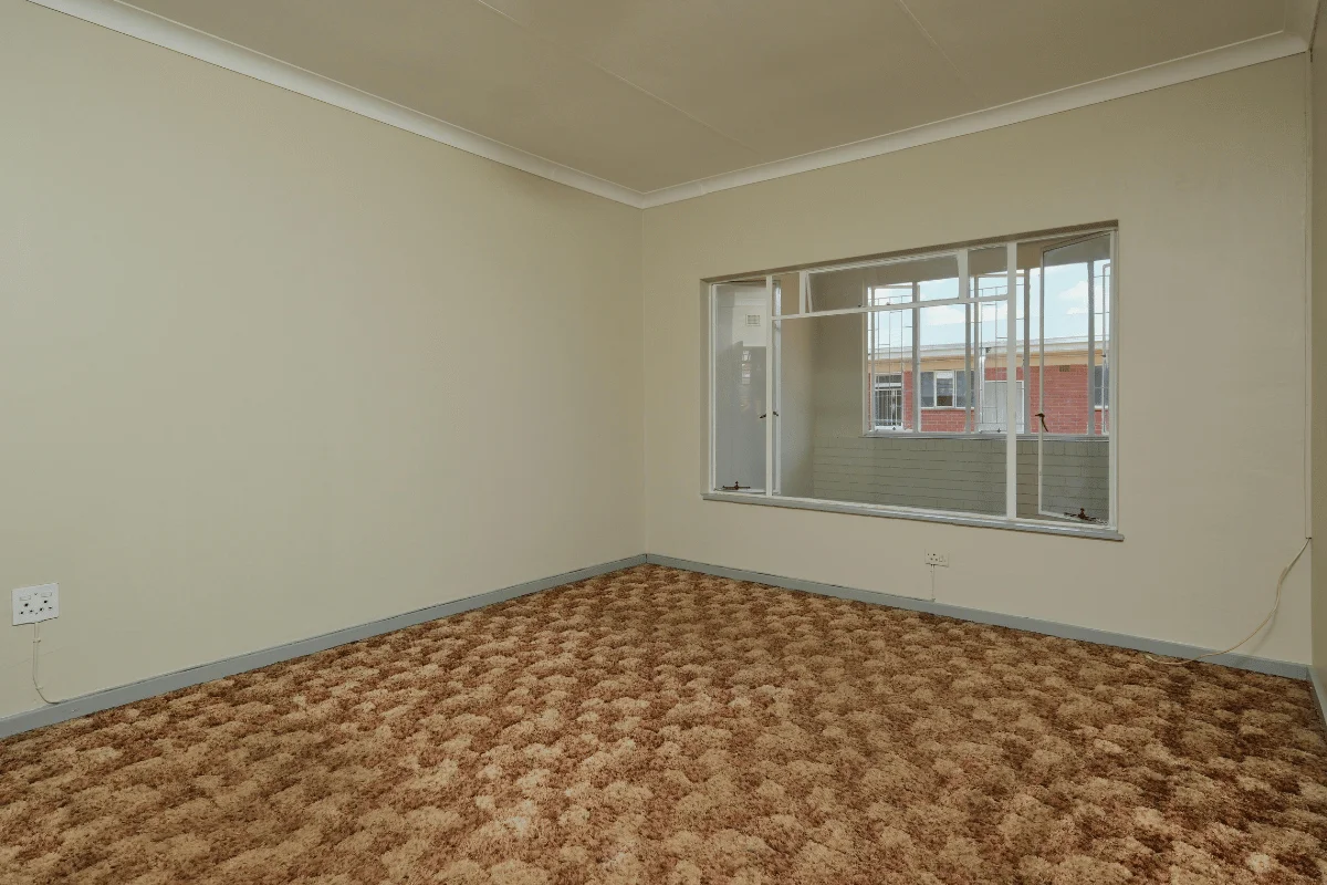 Empty room with beige walls and a large window showing an outdoor view. The floor is covered in brown, patterned carpet, creating a neutral, cozy feel.