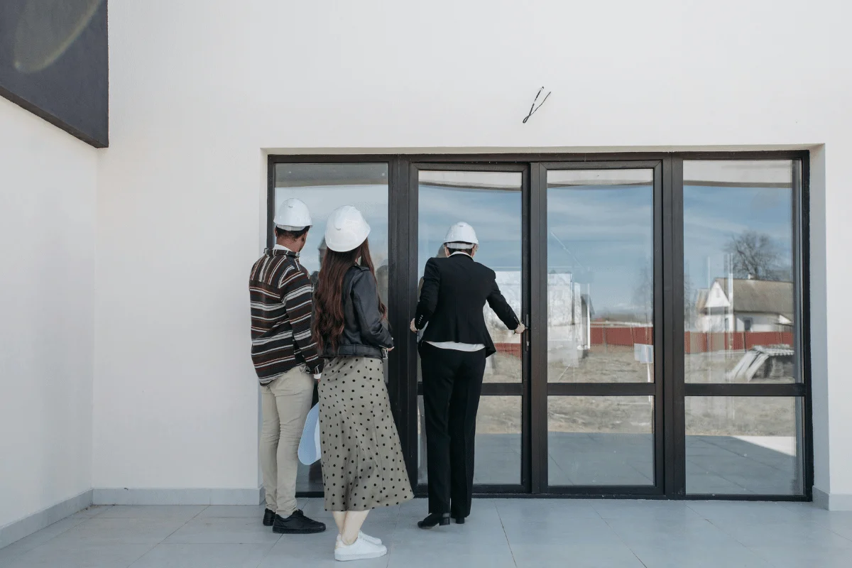 A couple and a real estate agent in hard hats stand in front of a glass door in a new construction building.