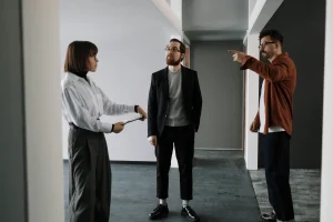 Three colleagues are in a hallway, engaged in a discussion. The woman on the left holds documents, the man on the right gestures emphatically, and the man in the center listens attentively.