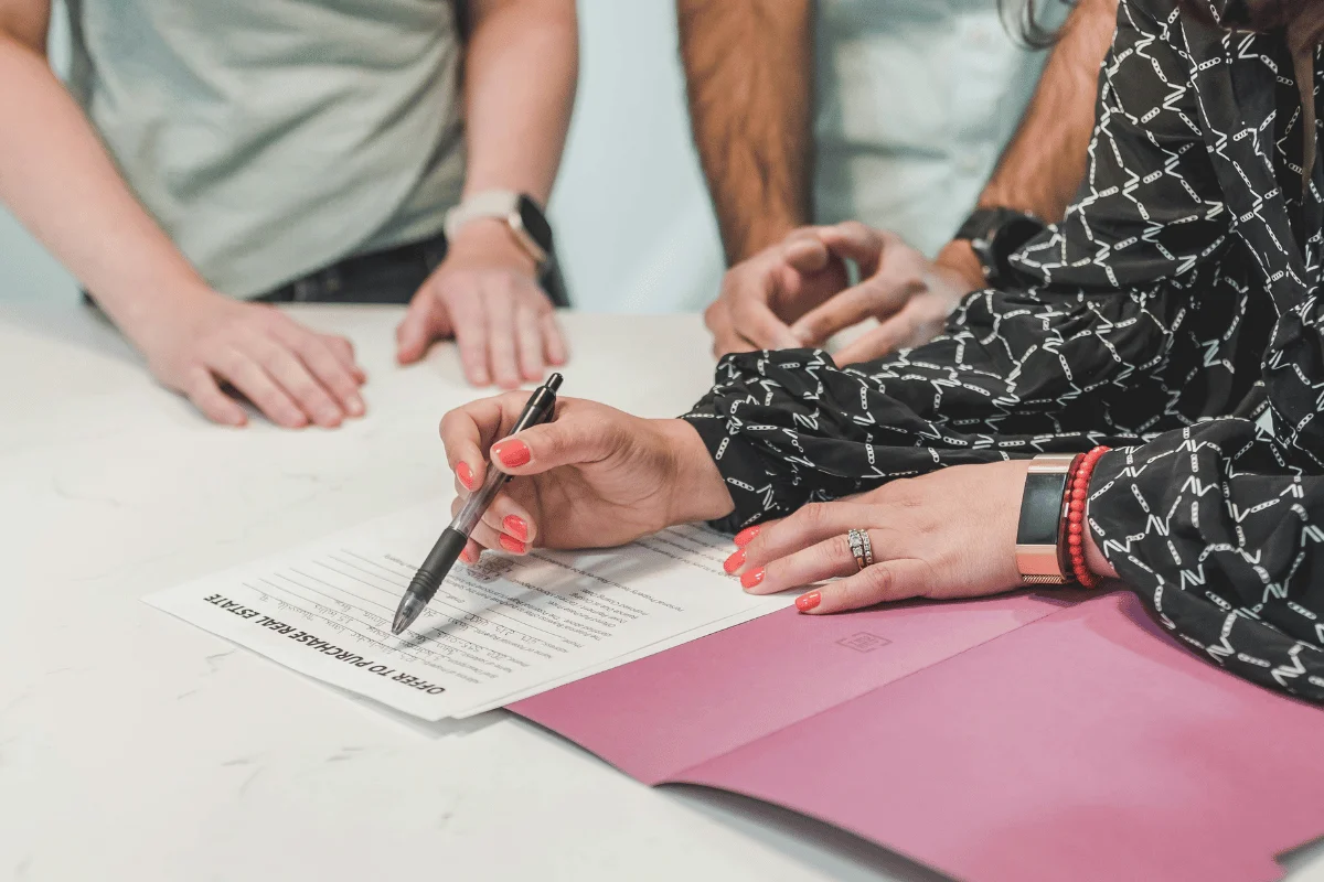 A close-up of a hand with red nails signing a real estate offer on a document.
