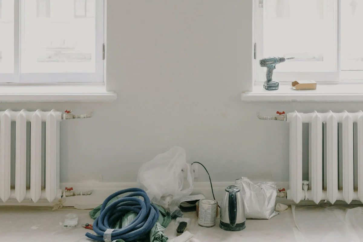 Room under renovation with two windows, radiators, a coiled blue hose, paint supplies, and a cordless drill on the window sill. The scene feels in-progress.