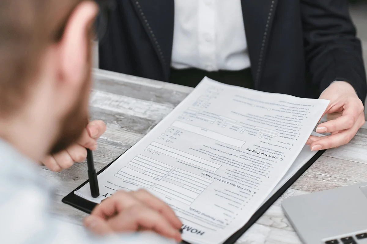 A person in a suit hands a printed document to another person holding a pen, suggesting a signing process. A laptop is partially visible on the table.