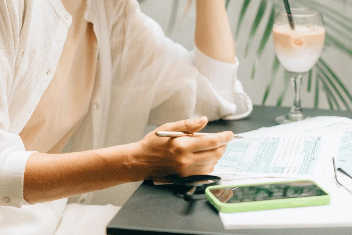 A person in a white shirt fills out forms at a table, holding a pen. A smartphone and glasses lie nearby.