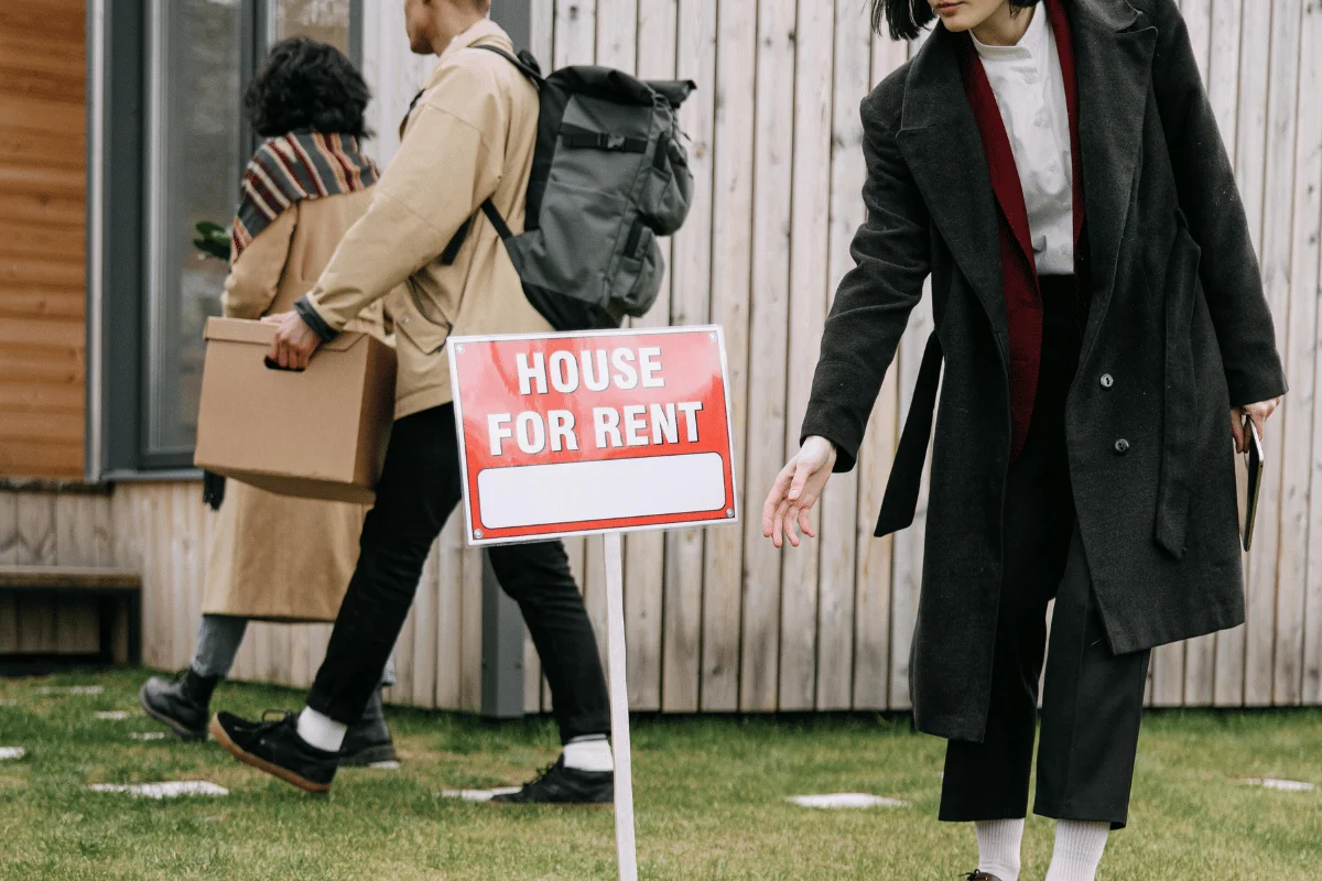 Woman adjusting a rental sign while others carry moving boxes.