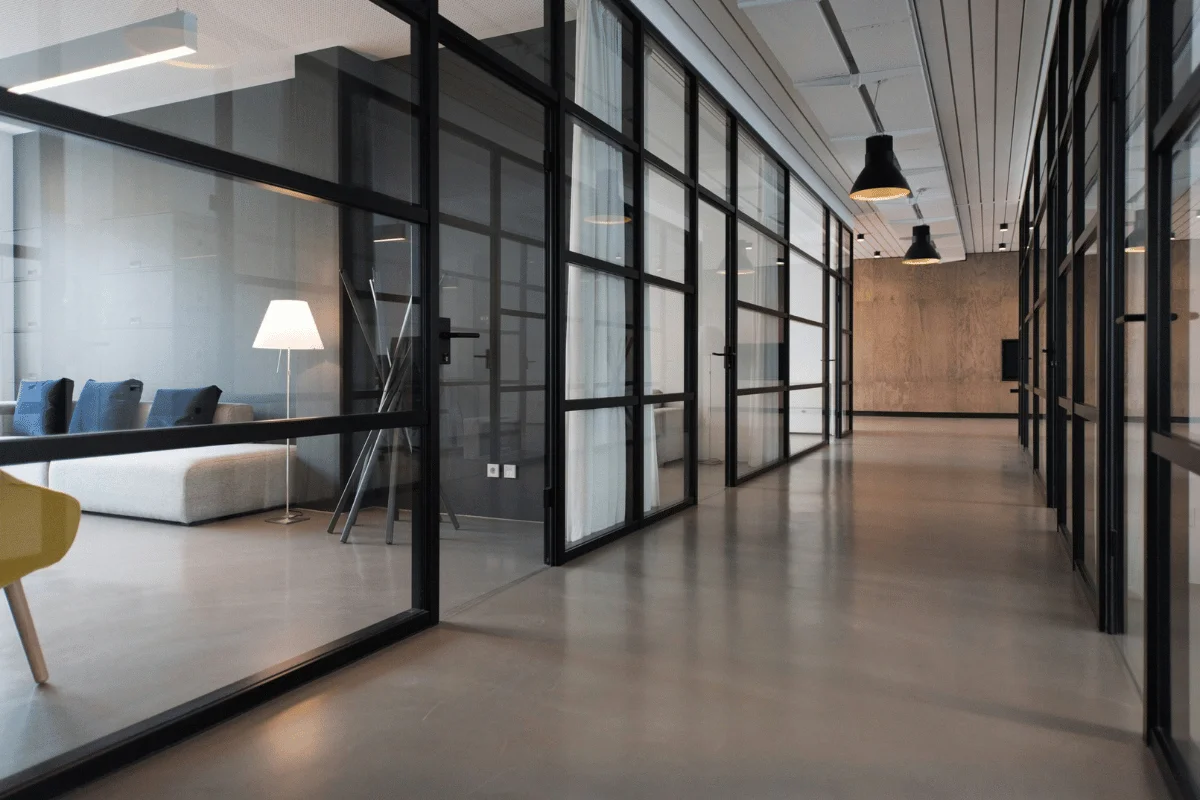 A modern office hallway with glass walls and doors, featuring a view into a cozy lounge area with a lamp and sofa.