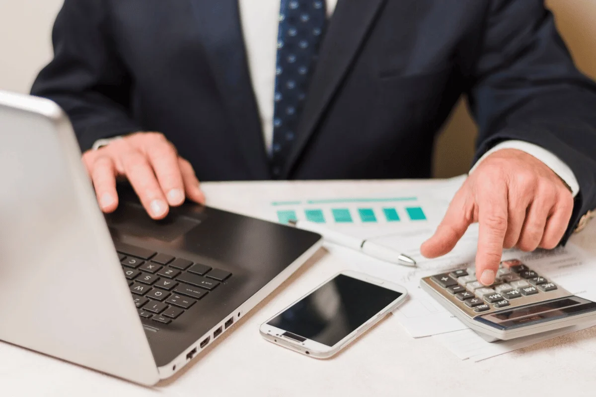 A man examines business financials using a calculator and laptop, surrounded by paperwork.