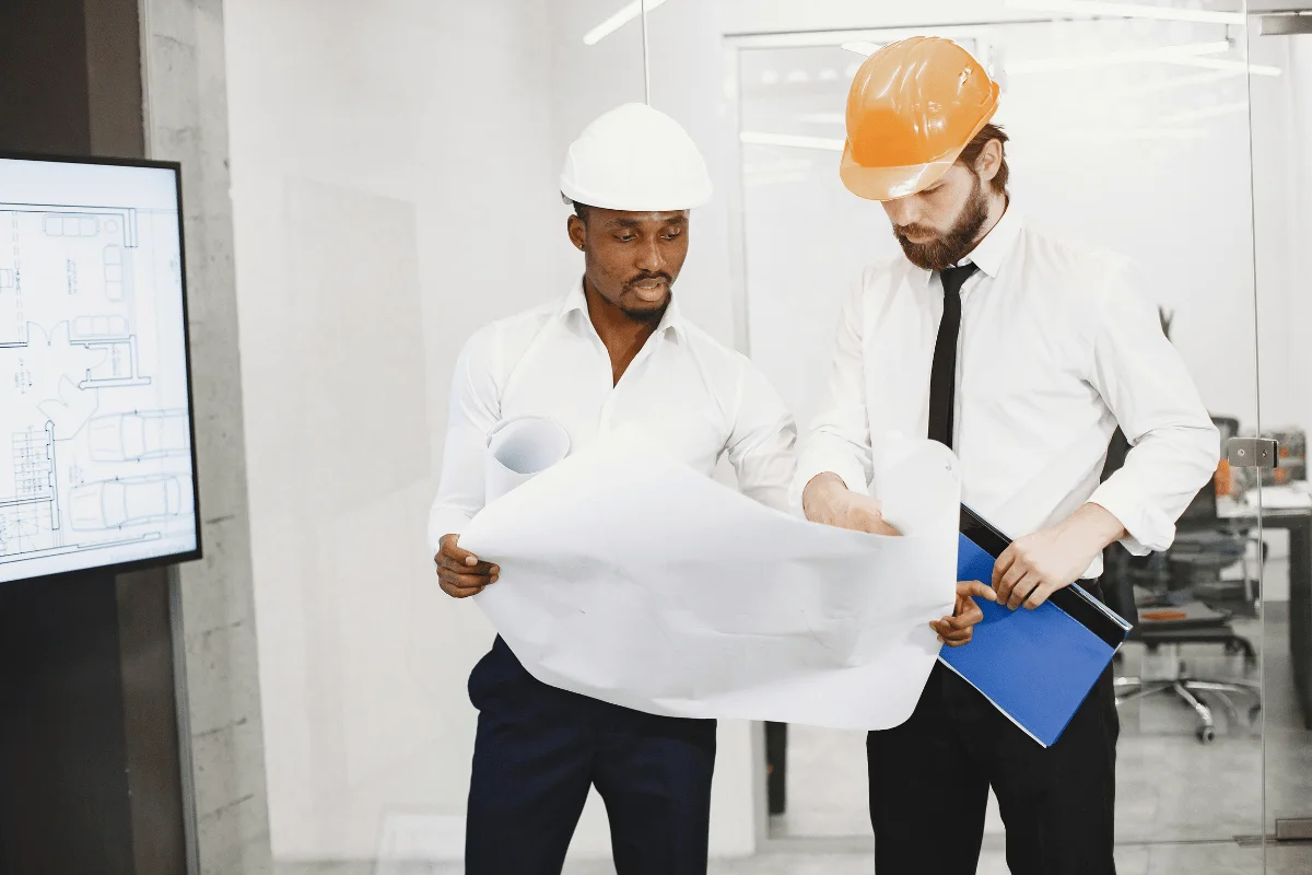 Two professionals in safety helmets discussing architectural plans in an office.