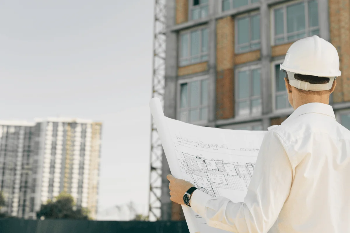 A construction manager in a hard hat examines blueprints at a building site, with a partially constructed apartment building in the background.