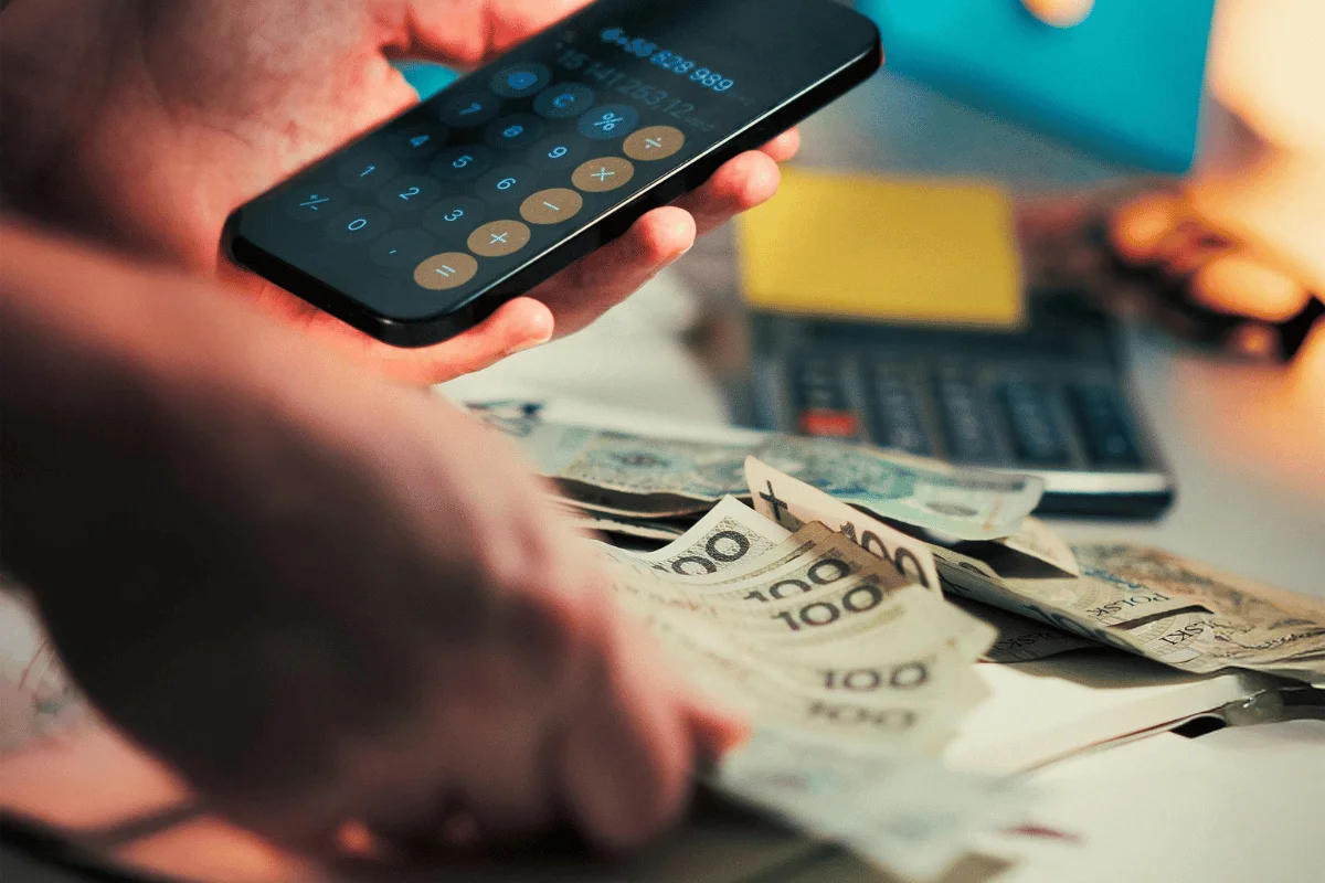A person calculates with a smartphone while handling assorted paper bills on a cluttered desk. Scene conveys financial planning and focus.