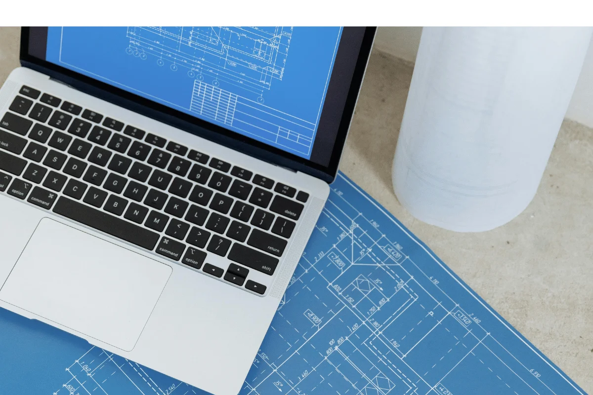 A laptop displaying blueprints on the screen with a rolled-up blueprint on the desk next to it.