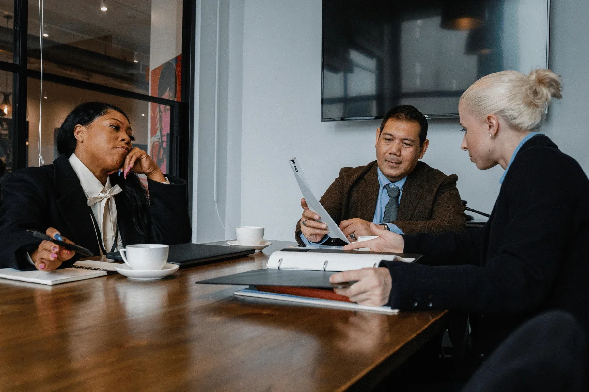 A group of professionals engaged in a discussion at a conference table, with notepads, a laptop, and coffee cups visible.