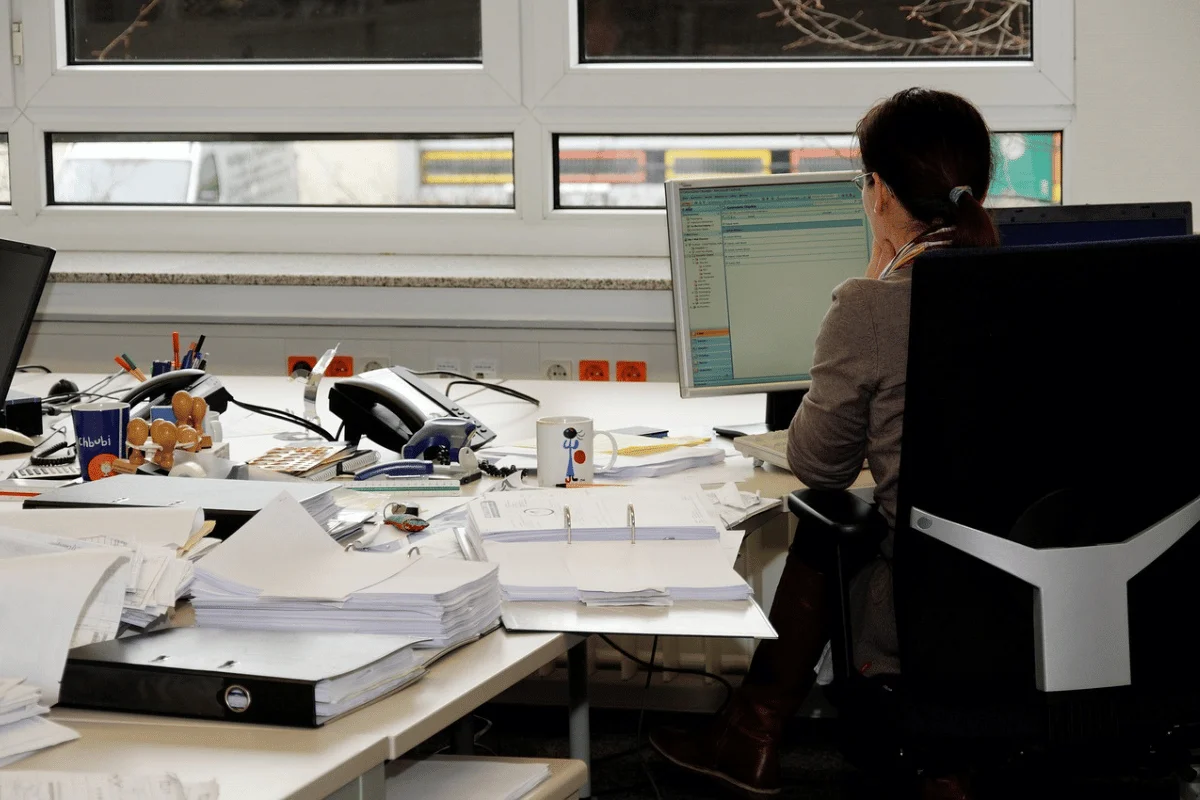 Office worker at computer surrounded by paperwork and files in a busy workspace.