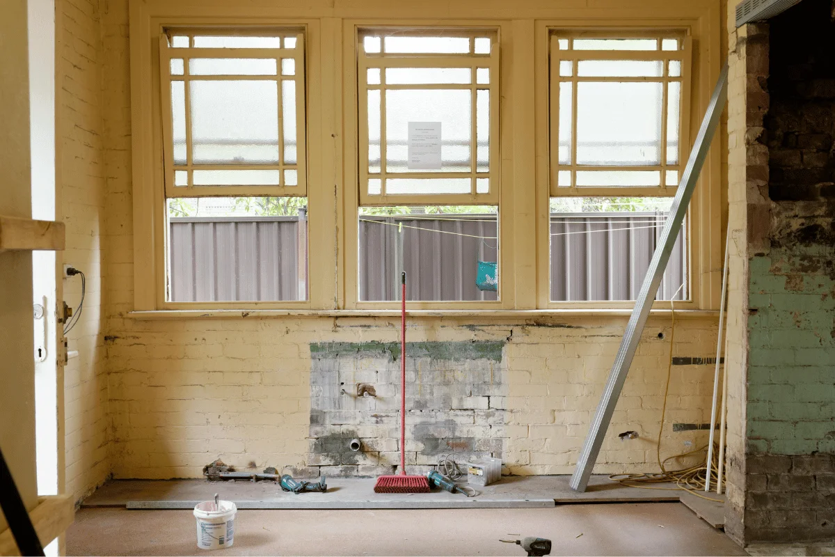 An empty, partially renovated room with three large windows, a broom, exposed pipes, and construction tools scattered. The atmosphere is unfinished and industrial.