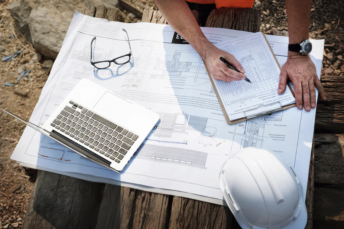 A construction worker is reviewing blueprints on a wooden table with a laptop and clipboard, writing notes.