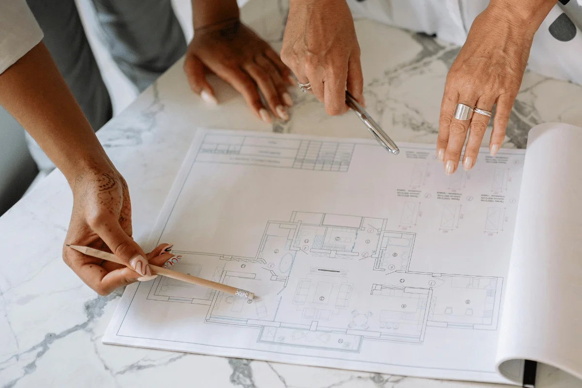 Two people review architectural blueprints on a marble table. One holds a pencil, the other points with a pen. The scene is collaborative and focused.