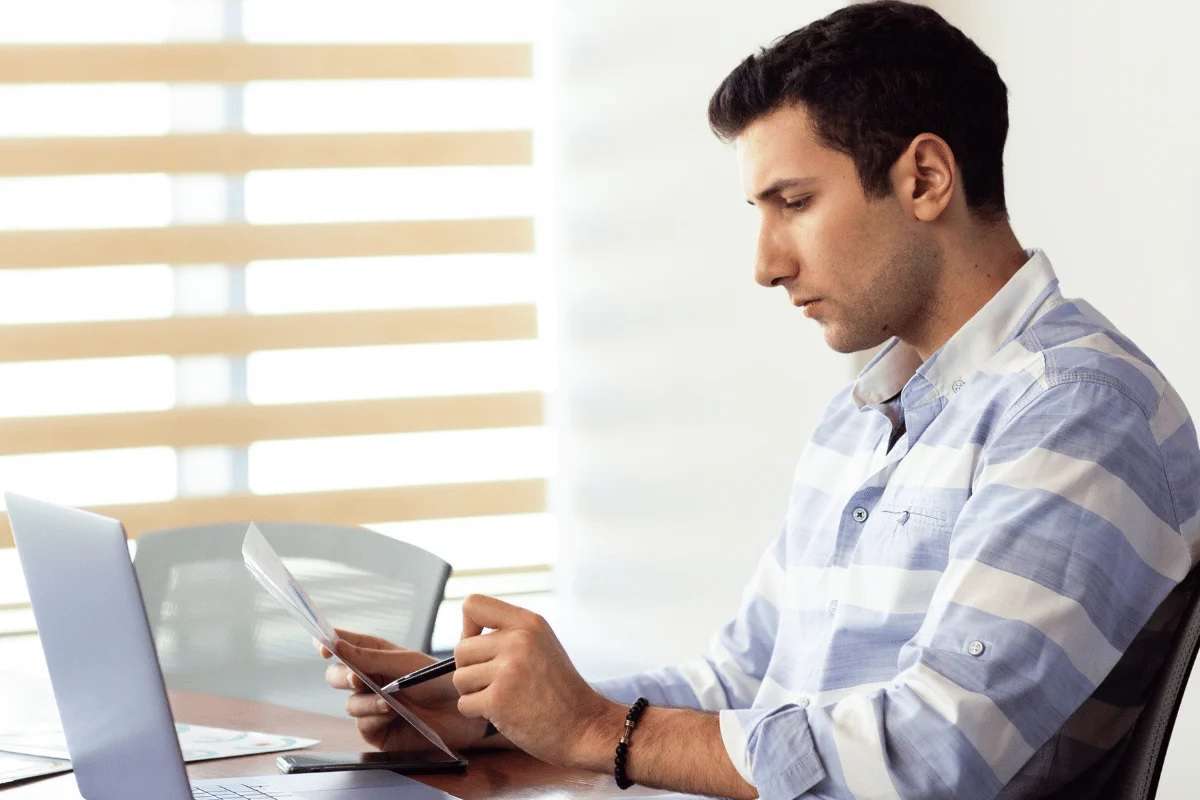 A focused man in a striped shirt is examining a document while seated at a desk with a laptop and a notebook.