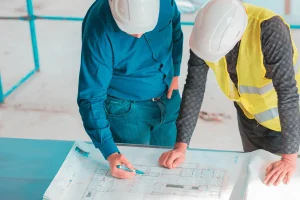 Two construction workers in hard hats review blueprints on a table. One wears a blue shirt, the other a yellow vest, focused on the plans.