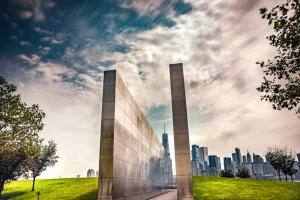 A striking memorial with two tall, parallel steel walls reflects a cloudy sky. In the distance, a city skyline is visible, surrounded by green grass and trees.