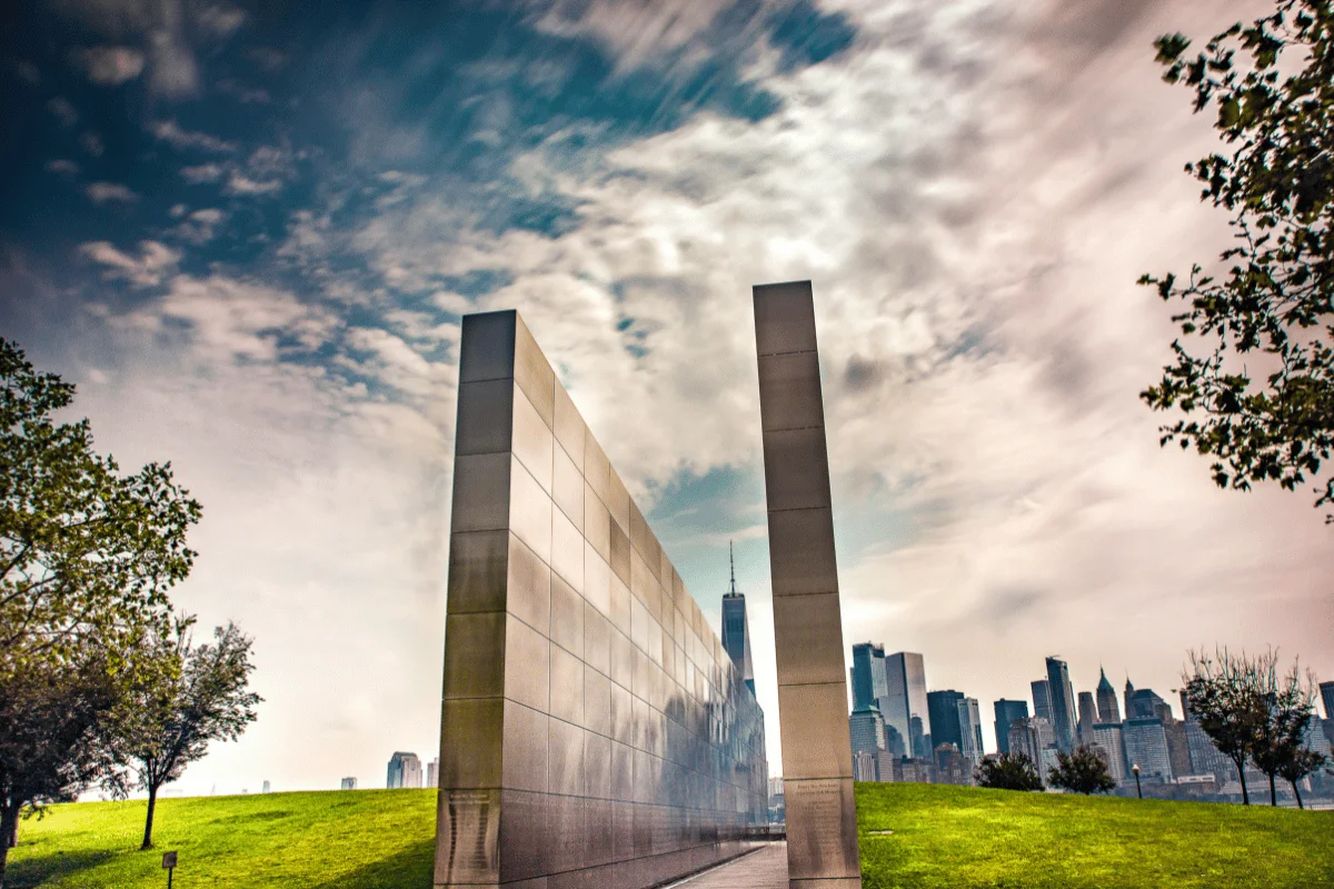 A striking memorial with two tall, parallel steel walls reflects a cloudy sky. In the distance, a city skyline is visible, surrounded by green grass and trees.