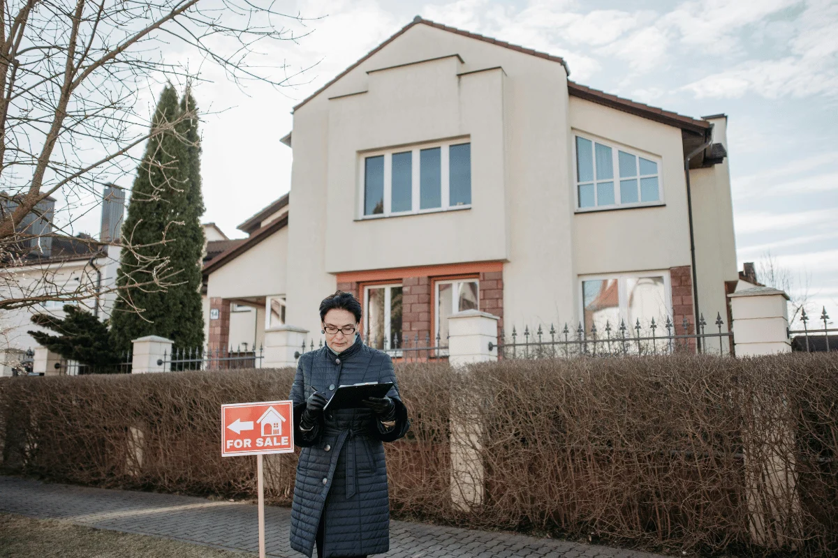 A woman in a coat holding a tablet outside a house with a "For Sale" sign in the front yard.
