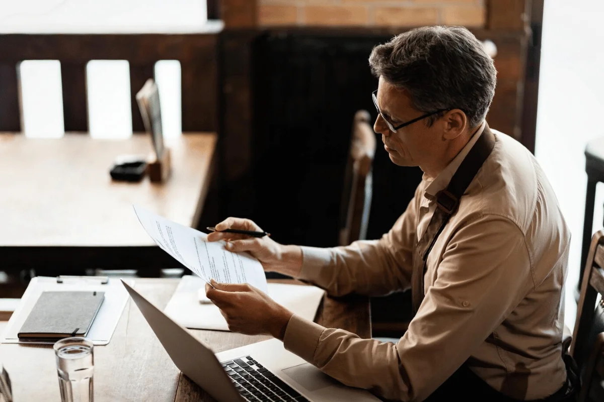 A professional man is working in a café, reading over documents and making notes while his laptop is open.
