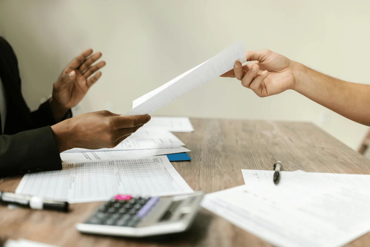 Two people exchanging financial documents across a desk covered with paperwork.