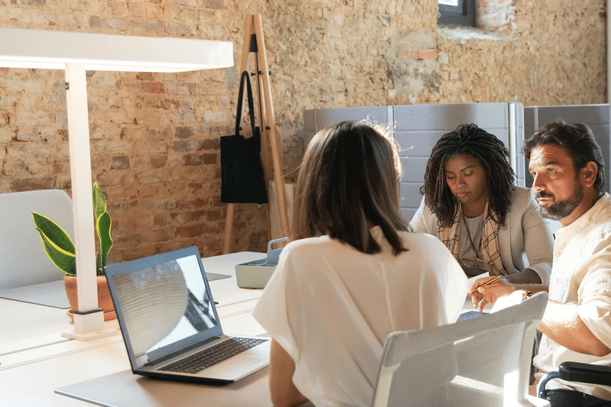 Office workers engaged in strategy session at shared desk with laptop and notes.