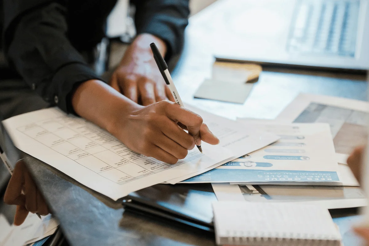 A close-up of a hand writing notes on a report amid documents, a notebook, and a laptop in a collaborative work environment.