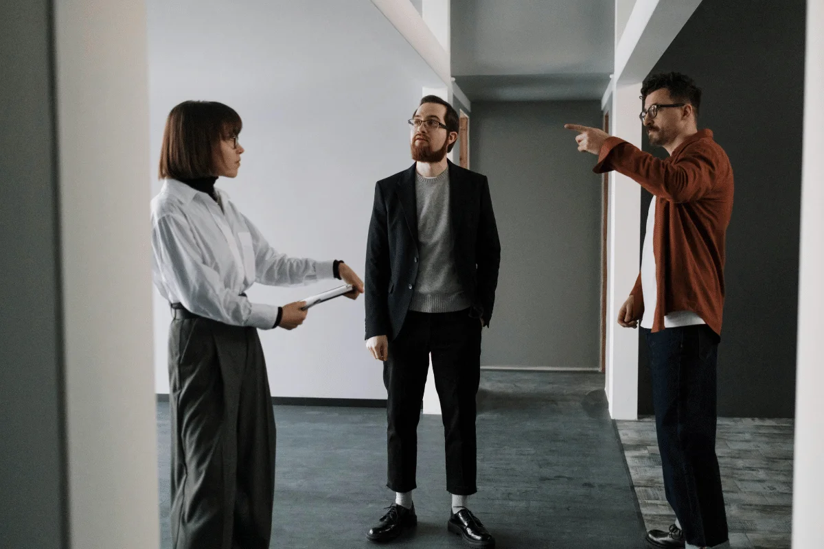 Three people stand in a modern empty room discussing property details while one person points toward a wall.