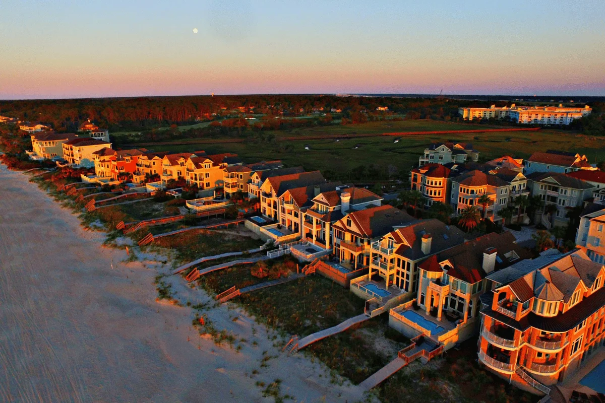 Aerial view of luxury beachfront houses at sunset, casting long shadows. The calm ocean and warm orange and pink skies create a serene atmosphere.
