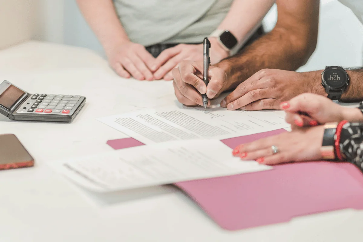 People signing financial documents on a desk with a calculator nearby.