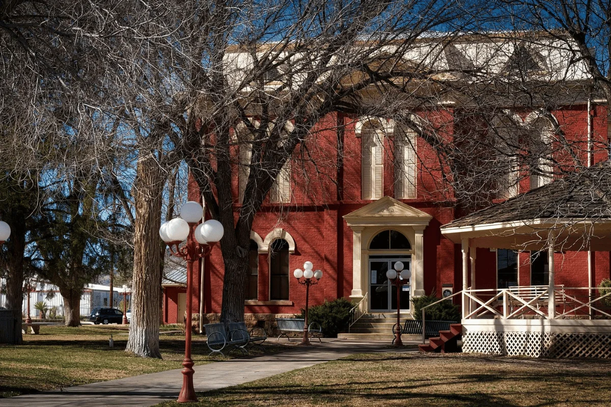 Historic red brick building framed by bare trees, with vintage street lamps and a gazebo in a grassy park setting.