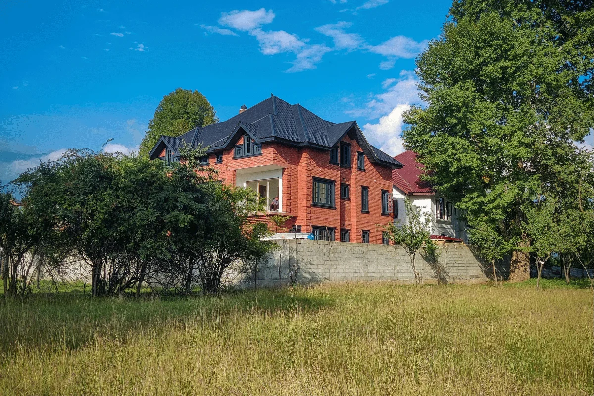 A charming red brick house with a black roof stands behind trees and a concrete wall, framed by a lush green lawn and a bright blue sky above.