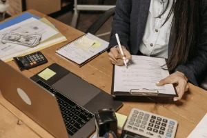 A person writing notes on a clipboard at a wooden desk, surrounded by a laptop, calculator, cash, and organized paperwork.