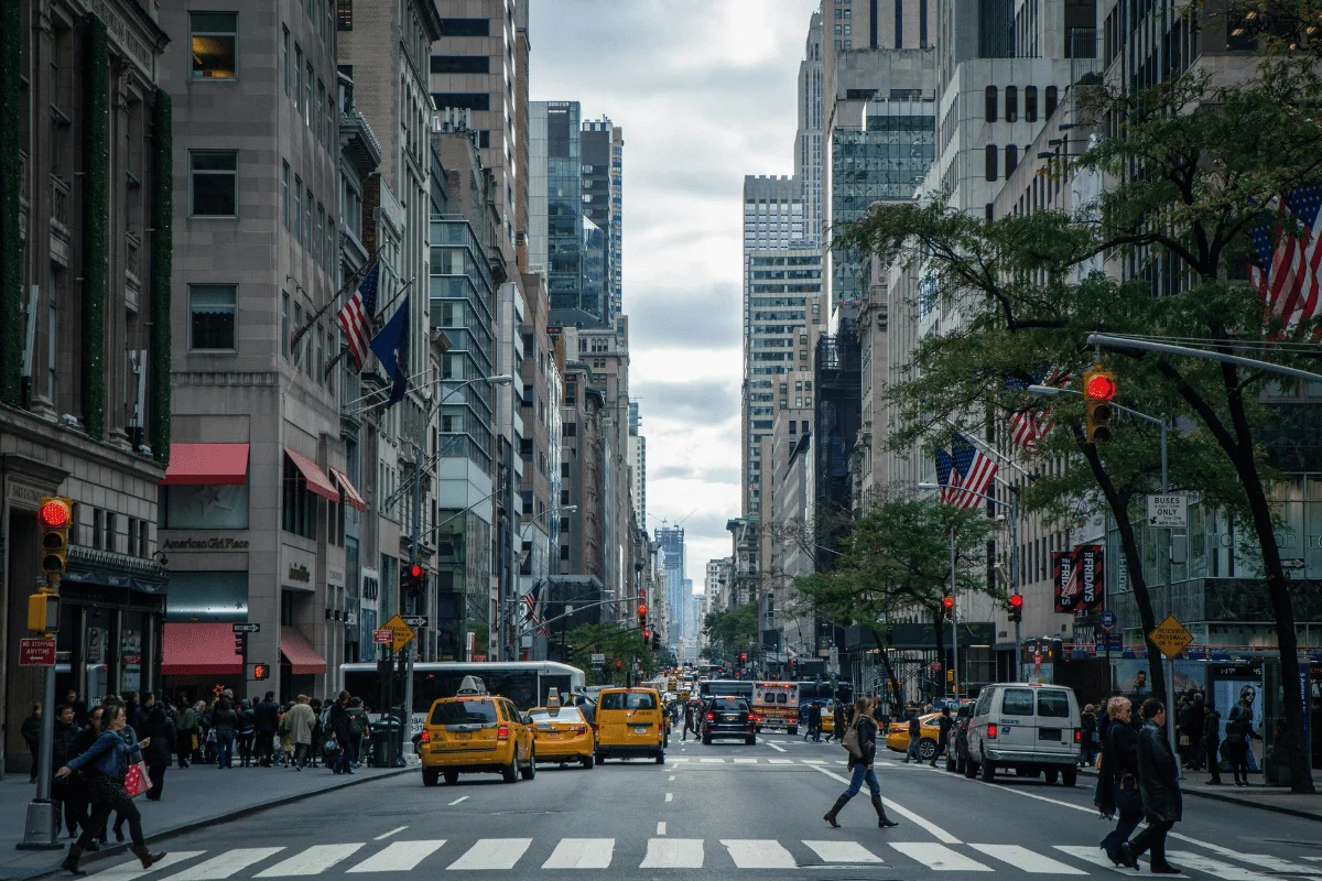 Bustling city street in New York with yellow taxis, pedestrians crossing, and tall buildings under a cloudy sky. U.S. flags line the avenue.