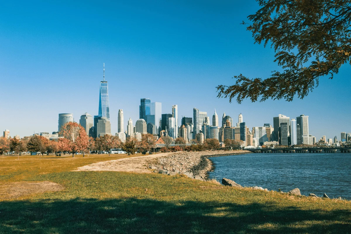 Panoramic view of a city skyline featuring modern skyscrapers under a clear blue sky. In the foreground, a grassy park borders a calm waterfront.