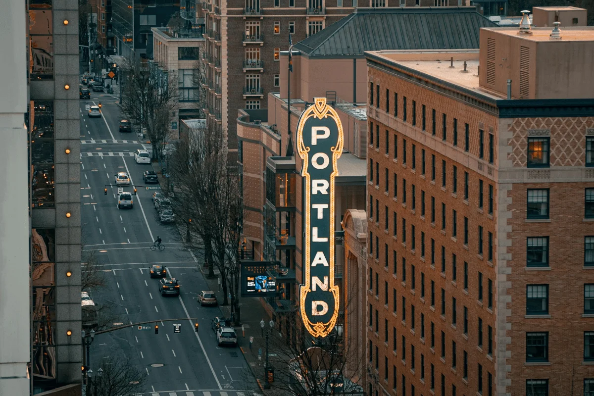 Aerial view of a city street lined with historic brick buildings. A large, illuminated "Portland" sign stands prominently, adding vibrant urban charm.