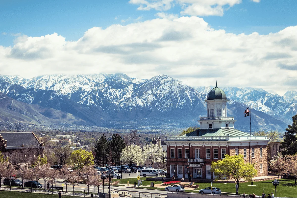 Historic brick building with a dome, lush trees, and cars in foreground. Majestic snow-capped mountains under a partly cloudy sky in the background.