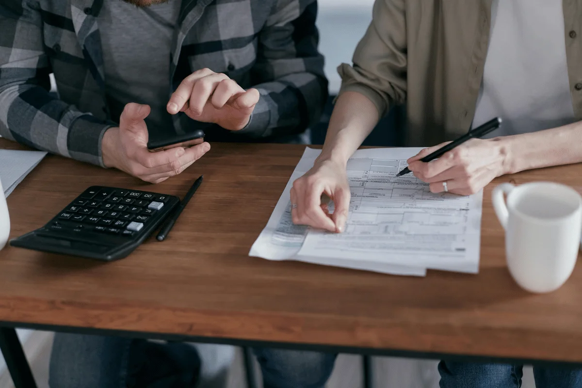 Two individuals collaborate at a wooden table, using a mobile phone and reviewing documents, with a calculator nearby and a coffee cup.