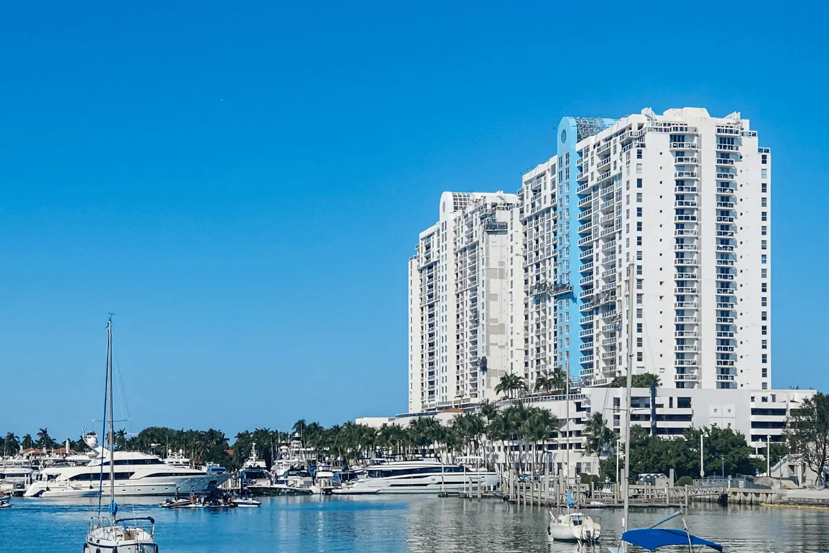 High-rise white buildings line a marina filled with yachts and sailboats, set against a clear blue sky.